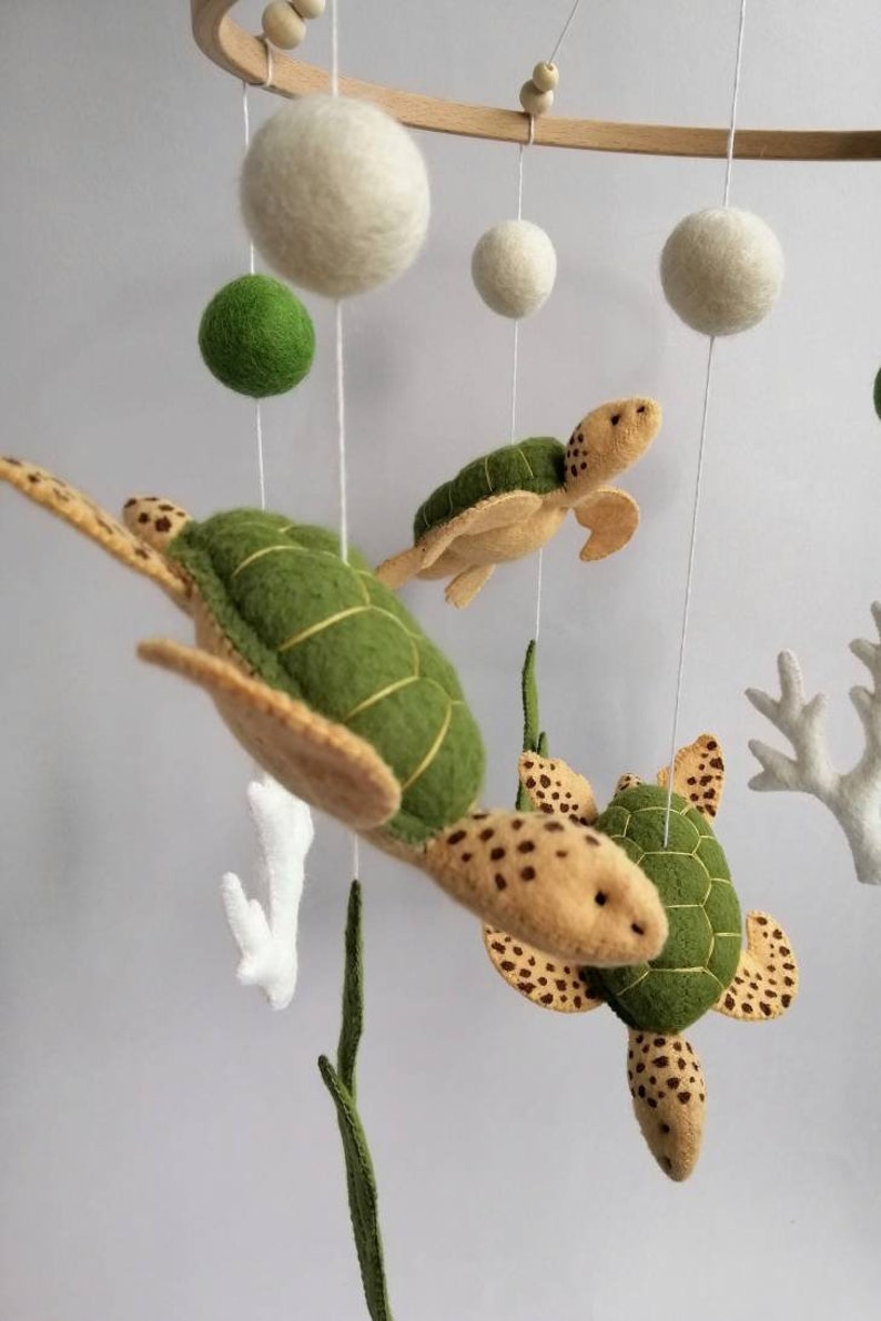 Puede incluir: Un m&oacute;vil de madera con decoraciones de fieltro de tortugas marinas y coral. El m&oacute;vil presenta tres tortugas verdes y marrones, coral blanco y hojas verdes. El m&oacute;vil est&aacute; suspendido de un anillo de madera.