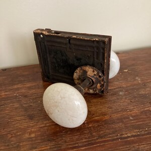 May include: Antique door hardware set featuring a black metal plate and two white ceramic oval doorknobs. The hardware shows signs of age and wear, with a rustic aesthetic. The set is designed for a door.