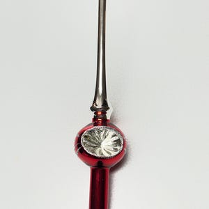 May include: A shiny red and silver glass tree topper featuring a long, slender silver spire extending upward from a central red sphere. The sphere has a reflective silver indented center and is decorated with thin, silver glittered line accents.