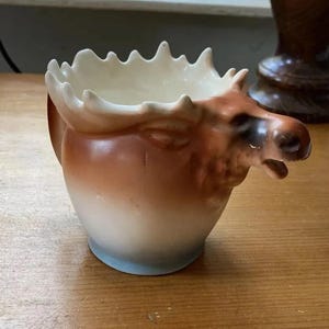 May include: A ceramic moose-head creamer with a brown and white gradient design. The moose head is detailed with a dark brown nose and antlers, and the rim of the creamer is scalloped.