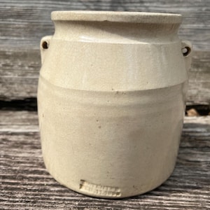 May include: A beige ceramic crock featuring a wide mouth, a thick rim, and two small side handles. The surface displays a fine crackle glaze pattern. Embossed text near the base reads PATENTED FEB 11 1890, indicating the historical manufacturing date.