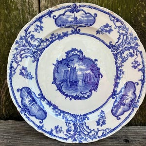 May include: A round ceramic plate featuring a white base with blue transferware patterns. The center depicts a nautical scene with sailboats on water, while the rim displays floral motifs and vignettes of windmills and ships.