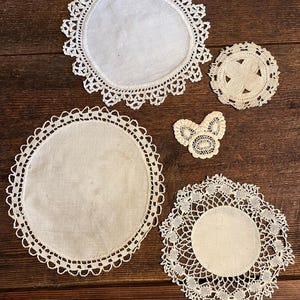 May include: A collection of five white, handmade doilies with intricate lace edges. The doilies vary in size and shape, including round and floral designs. They are arranged on a dark wooden surface.