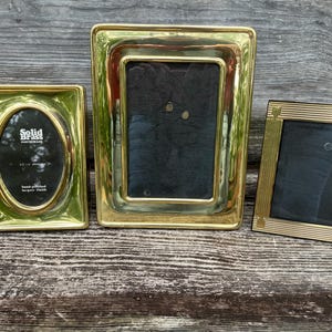 May include: Three brass picture frames of varying sizes. The frames have a polished finish and are rectangular and oval. The frames are empty, with dark backing. The frames are displayed on a weathered wooden surface.