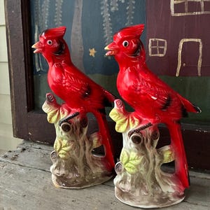 May include: Two matching ceramic cardinal figurines, each featuring a vibrant red bird with black facial markings perched atop a textured, light brown tree stump base with pale green leaves. The birds have open beaks and detailed plumage.