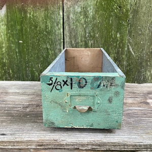 May include: A weathered, rectangular wooden box with a distressed teal paint finish. The box has a metal label holder and a small handle. The dimensions "5/8 x 10" are hand-painted in black on the side.