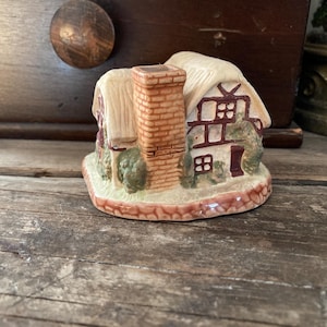 May include: A ceramic cottage figurine with a thatched roof, brick chimney, and detailed windows. The miniature house is painted in cream, brown, and green tones, and sits on a decorative base. A charming decorative piece.