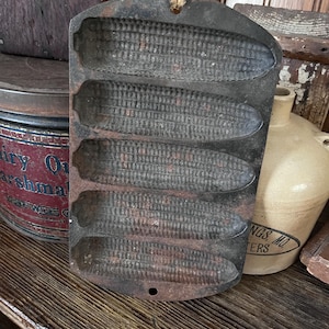 May include: A vintage cast iron cornbread mold with five corn-shaped cavities. The mold is dark gray with reddish-brown accents, showing signs of age and use. A jute string is attached for hanging. The mold is a kitchenware item.