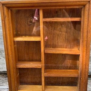 May include: A rectangular wooden display case with a glass front, featuring a natural brown wood grain finish. The interior is divided into two vertical sections with a total of five small horizontal shelves for organizing items.
