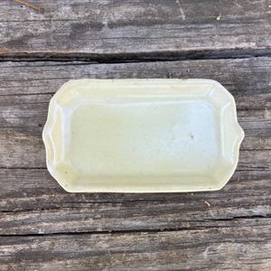 May include: A rectangular, light yellow ceramic tray with slightly raised edges and a smooth, glossy finish. The tray features a simple, clean design with rounded corners and a flat base, providing a functional surface for holding small items.