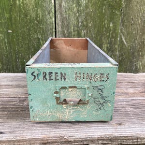 May include: A vintage, rectangular storage box with a distressed, turquoise paint finish. The box has a metal label holder and the words "SCREEN HINGES" are stenciled on the front. The box sits on a weathered wooden surface.