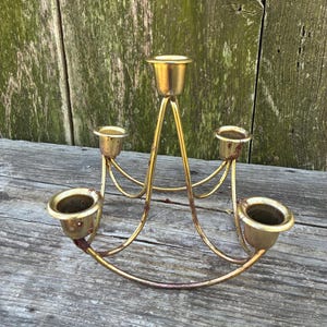 May include: A gold-toned metal candelabra featuring five candle holders arranged in a circular, tiered structure. The piece has thin, curved arms connecting the base to the elevated central holder, showing signs of wear and patina.