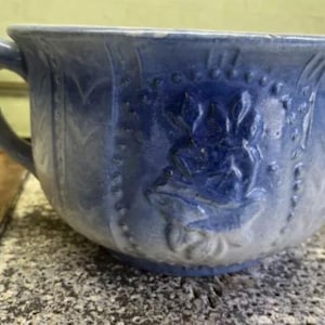 May include: A blue and white ceramic chamber pot with a floral design. The pot has a handle and a wide, rounded base.
