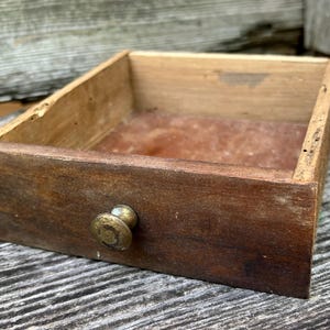 May include: A rectangular wooden drawer with a weathered brown finish and a single small brass-toned knob centered on the front panel. The interior base of the drawer features a reddish-brown surface, and the side edges show signs of natural wear.