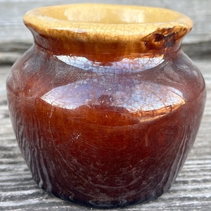 May include: A small, antique brown ceramic jar with a wide opening and a slightly flared rim. The jar has a glossy finish with visible crackling and a lighter tan-colored rim.