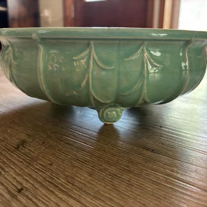 May include: A round, light green ceramic bowl featuring a scalloped, petal-like relief pattern along the exterior walls. The bowl is supported by three small, curled feet and has a smooth, glossy finish that reflects light across its curved surface.
