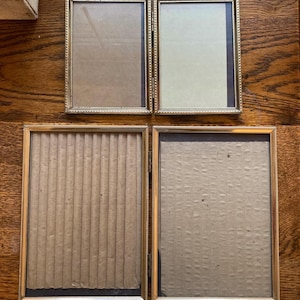 May include: Four rectangular gold-toned picture frames with ornate edges. Two frames have a light-colored backing, while the other two have a corrugated cardboard backing. The frames are empty, ready for photos or artwork. The frames are displayed on a wooden surface.