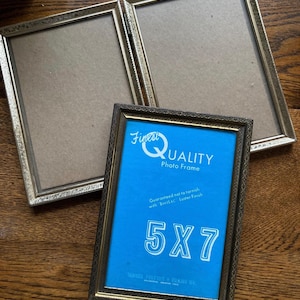 May include: Three gold-toned photo frames, two open and one closed, rest on a wooden surface. The closed frame features a blue backing with the text "Finest Quality Photo Frame" and "5X7". The frames have a decorative border.