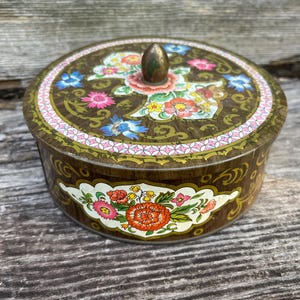May include: A round metal tin with a dark brown base color featuring a decorative floral pattern in shades of red, pink, yellow, and blue. The lid has a small, pointed brass-colored knob at the center and a pink and white geometric border.