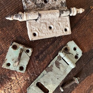 May include: An assortment of antique hardware components, including a decorative white hinge with ornate detailing, and two other metal pieces with signs of wear and age. The hardware is set against a rustic wooden surface.