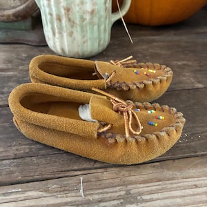 May include: A pair of tan suede moccasins featuring a whip-stitched toe seam, a decorative leather lace tie at the center, and small, multi-colored seed beads arranged in a simple pattern on the top of each shoe.