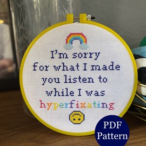 May include: A white embroidery hoop with a yellow frame. The embroidery features a rainbow, clouds, and the text "I'm sorry for what I made you listen to while I was hyperfixating". A yellow smiley face with a frown is below the text. The text "PDF Pattern" is in a blue circle below the hoop.