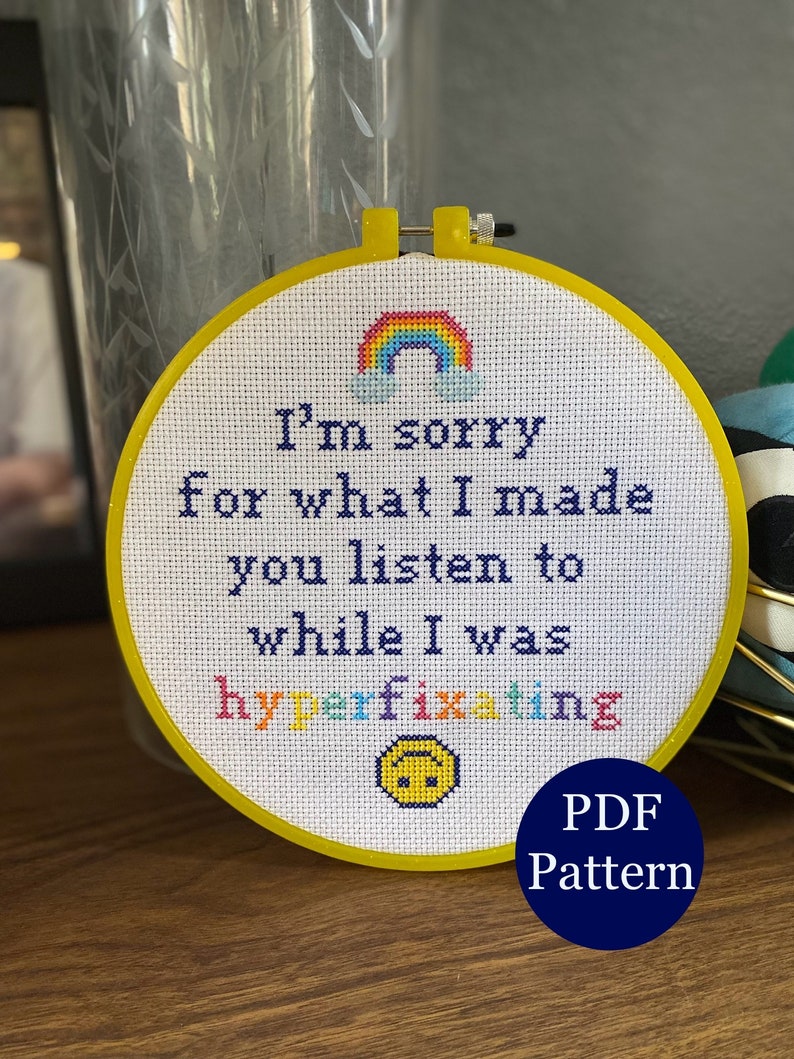 May include: A white embroidery hoop with a yellow frame. The embroidery features a rainbow, clouds, and the text "I'm sorry for what I made you listen to while I was hyperfixating". A yellow smiley face with a frown is below the text. The text "PDF Pattern" is in a blue circle below the hoop.