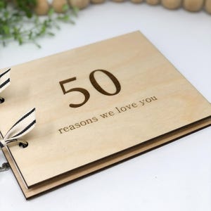 May include: A wooden photo album with a black and white ribbon. The cover is engraved with the number "50" and the text "reasons we love you".