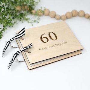 May include: A wooden photo album with a black and white ribbon closure. The front cover is engraved with the number "60" and the text "reasons we love you".