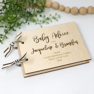 May include: A wooden baby advice book with a brown cover and black and white ribbon ties. The cover is engraved with the text "Baby Advice for Jacqueline & Brandon From Your Friends & Family That Love You! 2024".