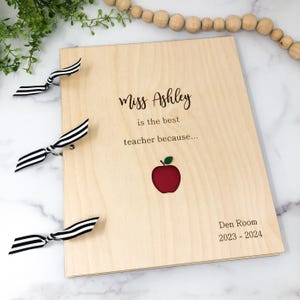 May include: A wooden teacher appreciation book with a red apple cutout and the text "Miss Ashley is the best teacher because... Den Room 2023 - 2024".