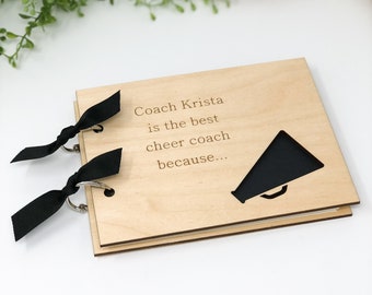 May include: A wooden scrapbook with a black megaphone cutout and the text "Coach Krista is the best cheer coach because..." on the front cover. The scrapbook has two black ribbon ties and silver rings.