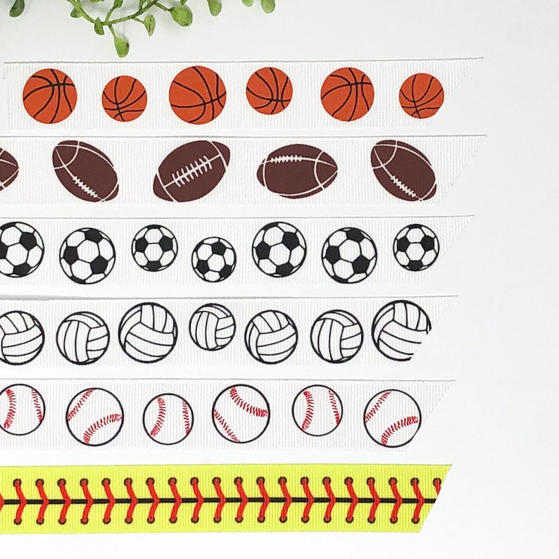 May include: Five white ribbons with different sports ball designs printed on them. The designs include basketballs, footballs, soccer balls, volleyballs, and baseballs. The last ribbon is yellow with red stitching, resembling a baseball.