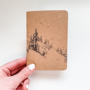 May include: A brown paper notebook with a black ink drawing of a mountain landscape with trees on the front cover.