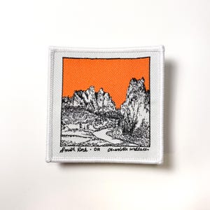 May include: White embroidered patch with an orange background featuring a black and white illustration of a mountain range with a river winding through it. The text "Smith Rock - OR Charlotte Wallace" is at the bottom of the patch.
