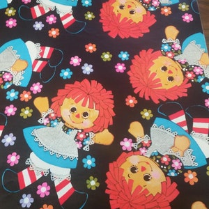 Könnte beinhalten: Einpackpapier mit einem sich wiederholenden Muster aus rothaarigen Puppen, die blaue Kleider und weiße Spitzen-Schürzen tragen. Die Puppen befinden sich auf einem schwarzen Hintergrund mit bunten Blumen.