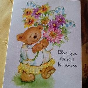 May include: Greeting card featuring a teddy bear holding a bouquet of colorful flowers. The bear wears a white dress with a yellow ruffled skirt. The text "Bless You for Your Kindness" is printed on the card.