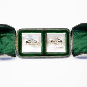 May include: An open black leather jewelry box with a green velvet interior. The box contains two silver cufflinks and two gold rings. The cufflinks are in a rectangular compartment, and the rings are in the side compartments.