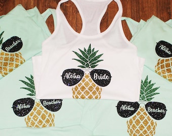 May include: Set of seven light green tank tops with a pineapple design and the text "Aloha Beaches". One white tank top has a pineapple design and the text "Aloha Bride".