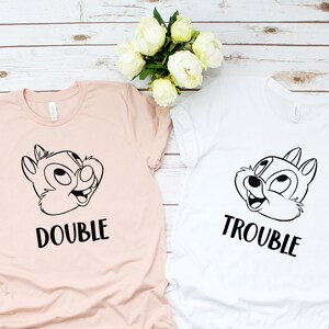 Puede incluir: Dos camisetas con un diseño de ardilla de dibujos animados. Una camiseta es rosa y dice "Double Trouble" y la otra es blanca y dice "Trouble".