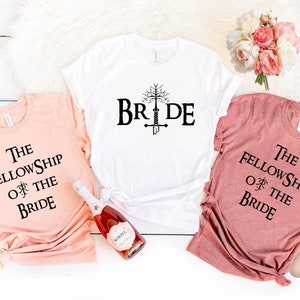 May include: Three t-shirts with a Lord of the Rings theme for a bridal party. The white shirt says "Bride" with a tree and sword graphic. The peach shirt says "The Fellowship of the Bride". The pink shirt says "The Fellowship of the Bride".