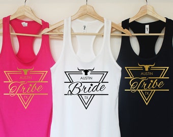 Puede incluir: Tres camisetas de tirantes con un diseño geométrico en oro y negro que presenta una cabeza de toro y el texto "Austin Tribe TX". Las camisetas de tirantes son rosa, blanca y negra.