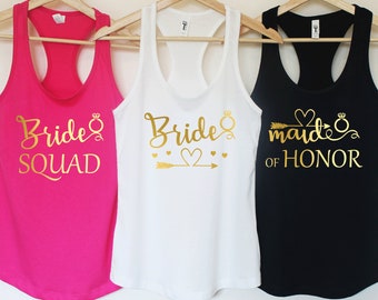 May include: Three tank tops with gold lettering. The pink tank top says "Bride Squad", the white tank top says "Bride", and the black tank top says "Maid of Honor".