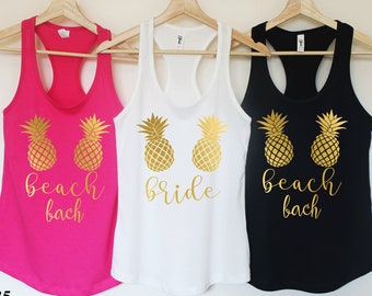 May include: Three tank tops with a pineapple design and text. The tank tops are pink, white, and black. The text on the pink tank top says "beach bach". The text on the white tank top says "bride". The text on the black tank top says "beach bach".