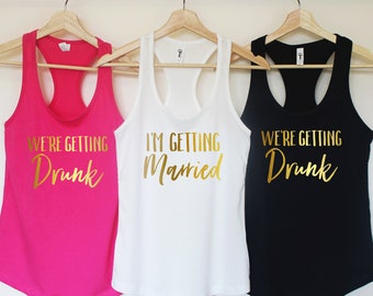 May include: Three tank tops with gold lettering. The pink tank top says "We're Getting Drunk". The white tank top says "I'm Getting Married". The black tank top says "We're Getting Drunk".