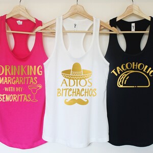 Puede incluir: Tres camisetas sin mangas con letras y gr&aacute;ficos dorados. La camiseta rosa dice "Drinking Margaritas - With My Senoritas" y tiene un gr&aacute;fico de un vaso de margarita. La camiseta blanca dice "Adios Bitchachos" y tiene un gr&aacute;fico de un sombrero. La camiseta negra dice "Tacoholic" y tiene un gr&aacute;fico de un taco.