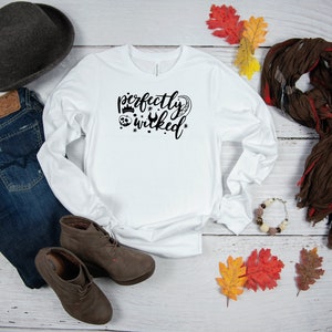 May include: White long-sleeve shirt with the text "perfectly wicked" in black lettering. The shirt is styled with jeans, boots, a hat, a scarf, and fall leaves.