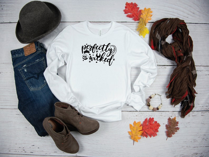 May include: White long-sleeve shirt with the text "perfectly wicked" in black lettering. The shirt is styled with jeans, boots, a hat, a scarf, and fall leaves.