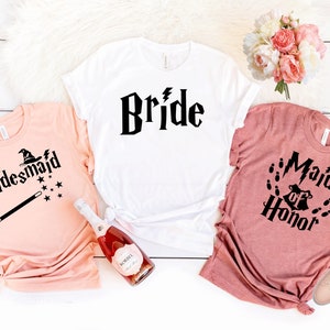 May include: Three t-shirts with a Harry Potter theme for a wedding party. The white shirt says "Bride" in a black font. The peach shirt says "Bridesmaid" with a black witch hat and magic wand. The pink shirt says "Maid of Honor" with a black Hogwarts crest and footprints.