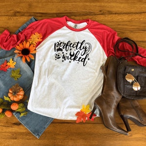May include: A white and red baseball tee shirt with the text "perfectly wicked" in black. The shirt is styled with a pair of blue jeans, a brown leather bag, and brown leather boots.